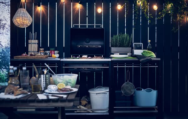 Guirlande lumineuse suspendue au-dessus d’une cuisine extérieure composée de trois éléments, dont un barbecue, adossés à une barrière en bois noire.