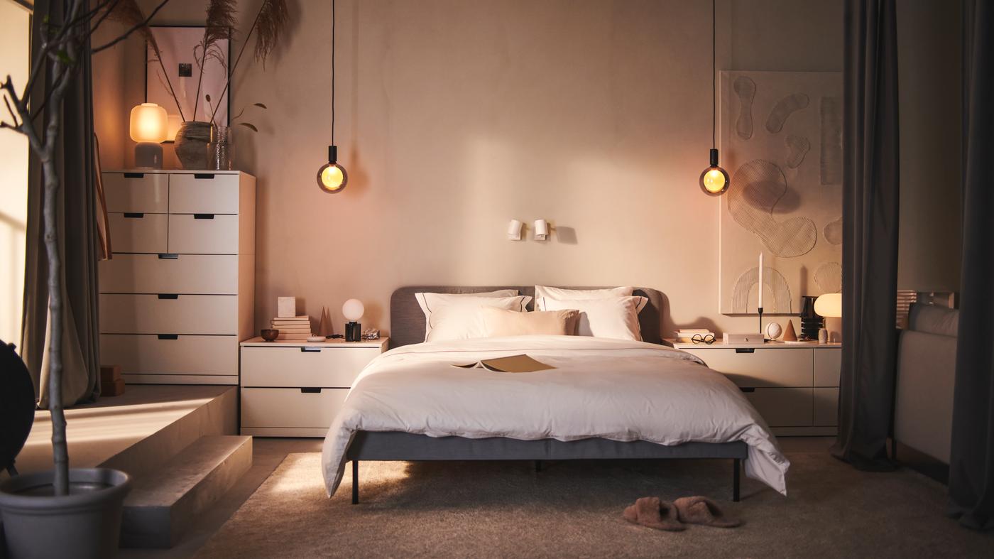 Habitación con cama doble o cama matrimonial junto a cómodas blancas y dós lámparas de techo con ampolletas redondas.