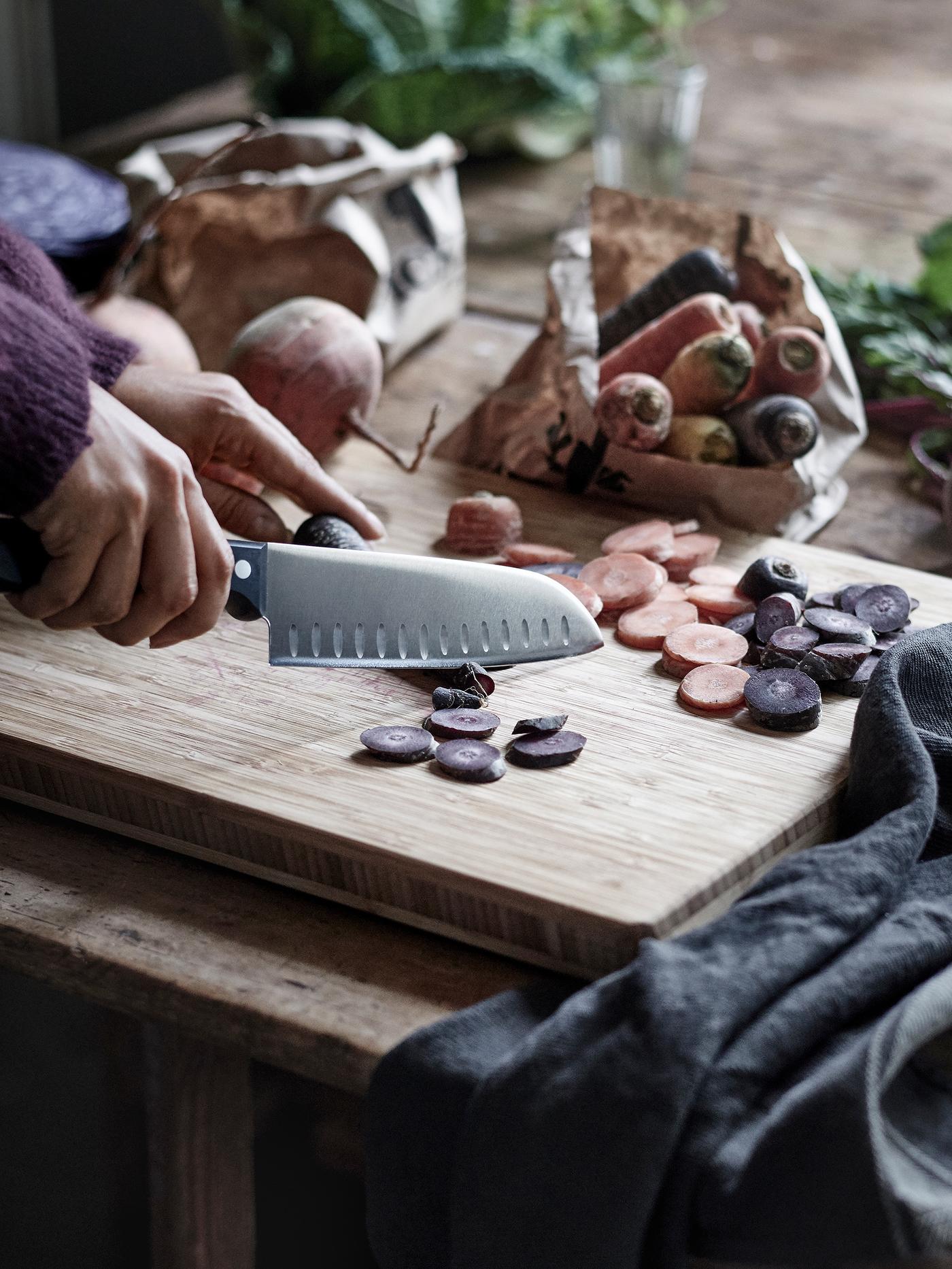 Hands chopping carrots on a wooden chopping board using a utility knife with a black handle.