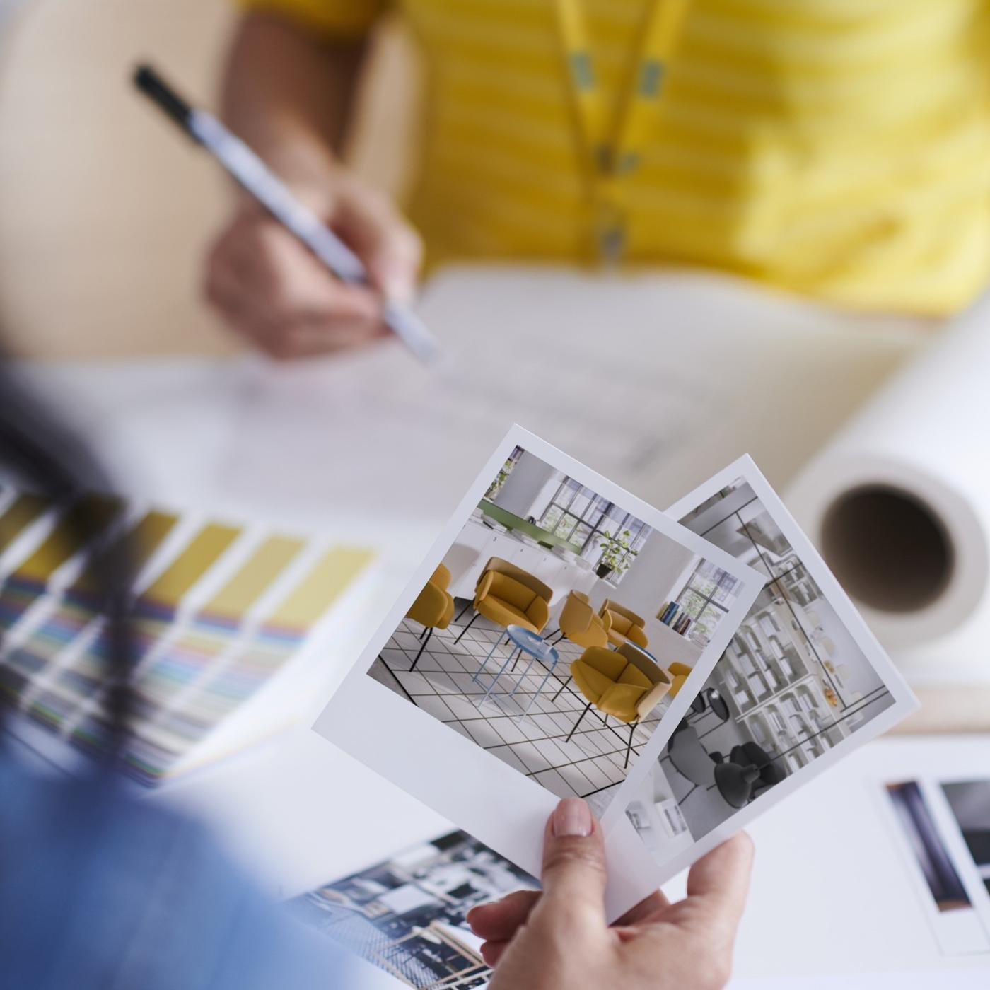 Hands holding printed interior design images over a desk with colour swatches, drawings, and design materials, showing a planning or design process.