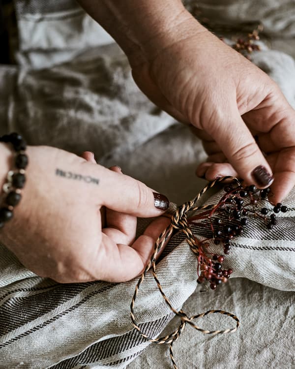 Hands tying a decorative knot made from natural and black coloured twine and plum coloured wild berries round a napkin.