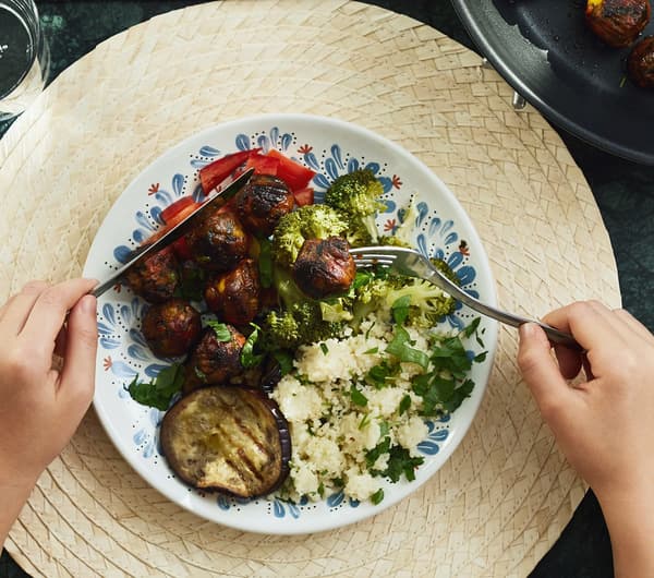 Hands with a knife and fork held over a plate of vegan food, including IKEA ALLEMANSRÄTTEN veggie balls, on a placemat.