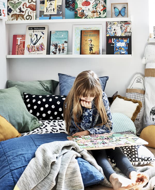 Hania liest auf ihrem Bett mit bunter Bettwäsche. Hinter ihr ist MOSSLANDA Bilderleiste weiss mit bunten Büchern darauf zu sehen.