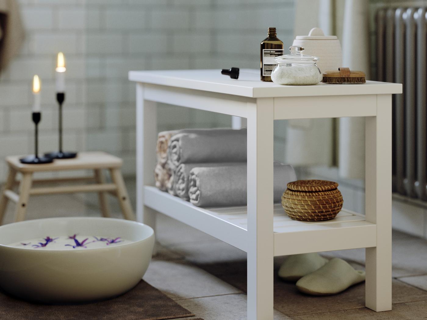 HEMNES bænk står i badeværelse med sterinlys, fodbad og håndklæder ligger på hylden. 