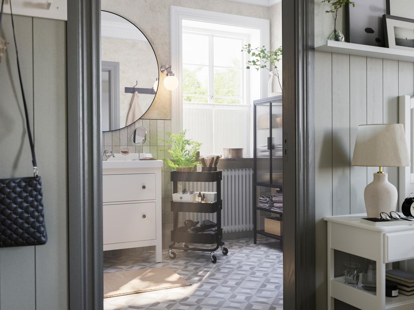 큰 창이 있는 회색 욕실에 HEMNES 헴네스 화이트 세면대와 RÅSKOG 로스코그 블랙 카트, MOSSJÖN 모셴 유리도어수납장이 있는 모습.