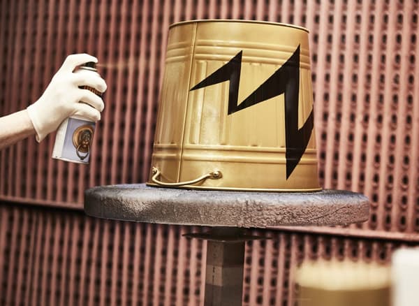 Hier wird KNODD Tonne mit Deckel grau gerade mit goldfarbener Sprühfarbe neu lackiert. In der Mitte ist ein schwarzer Blitz als Dekoration zu erkennen.
