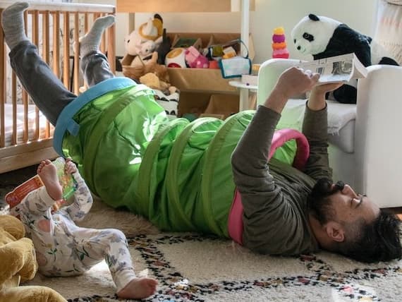 Hombre dentro de una cuncuna de juegos para niños recostado en el piso sobre una alfombra de color blanco con gris, a su lado un bebé en la misma posición que el y peluches.
