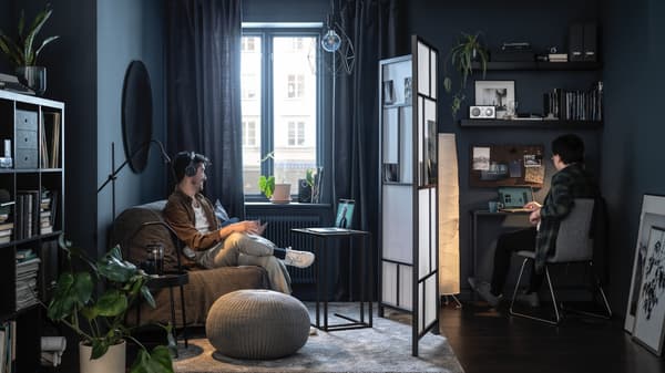 Hombre sentado escuchando música en un sillón gris LYCKSELE LÖVÅS viendo un programa en su notebook y paralelo hay otro hombre trabajando en su computador sentado en una silla y apoyado en un escritorio.