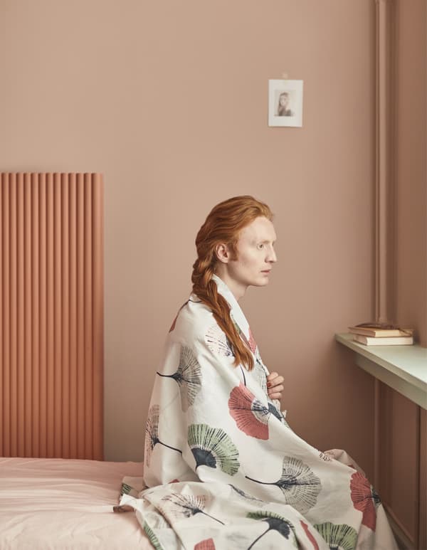 Homme à la chevelure rousse assis sur un lit avec un tissu drapé sur les épaules.