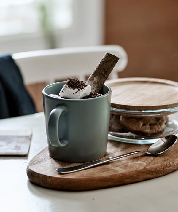 Hot chocolate with marshmallows in a grey-blue mug served on a chopping board with cookies in a glass jar with bamboo lid.