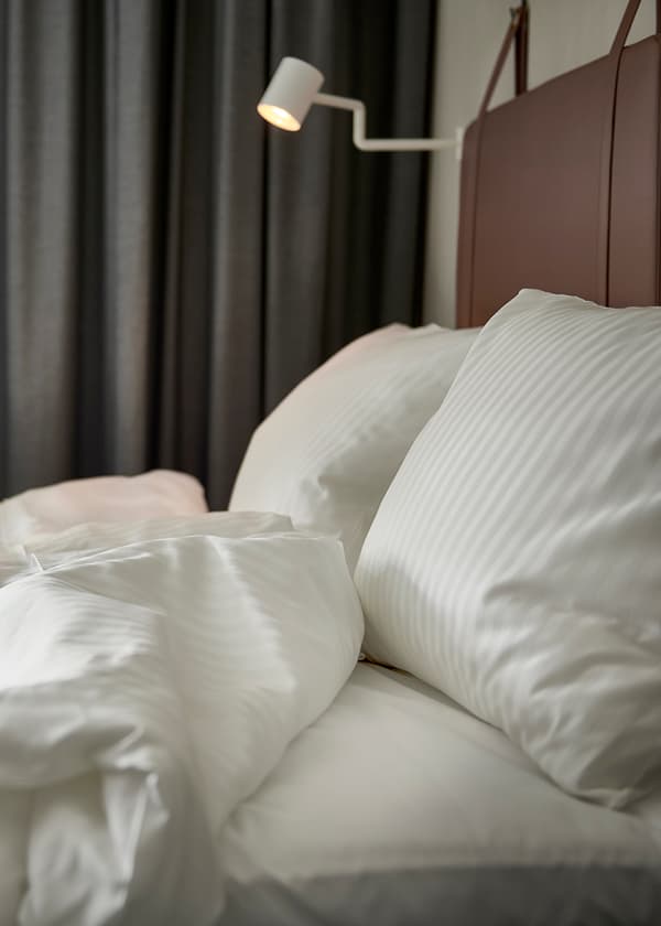 Hotellsäng med vita lakan och kuddar, samt en väggmonterad läslampa ovanför.