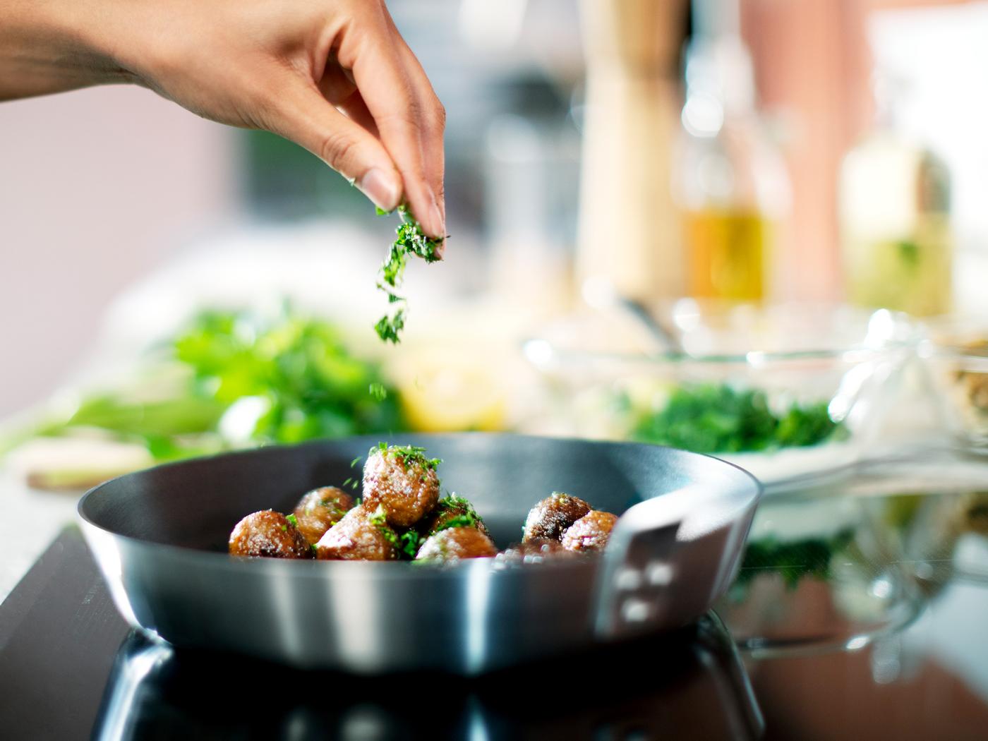HUVUDROLL plant balls being fried in a black frying pan on an induction hub. A hand is sprinkling chopped parsley over them.