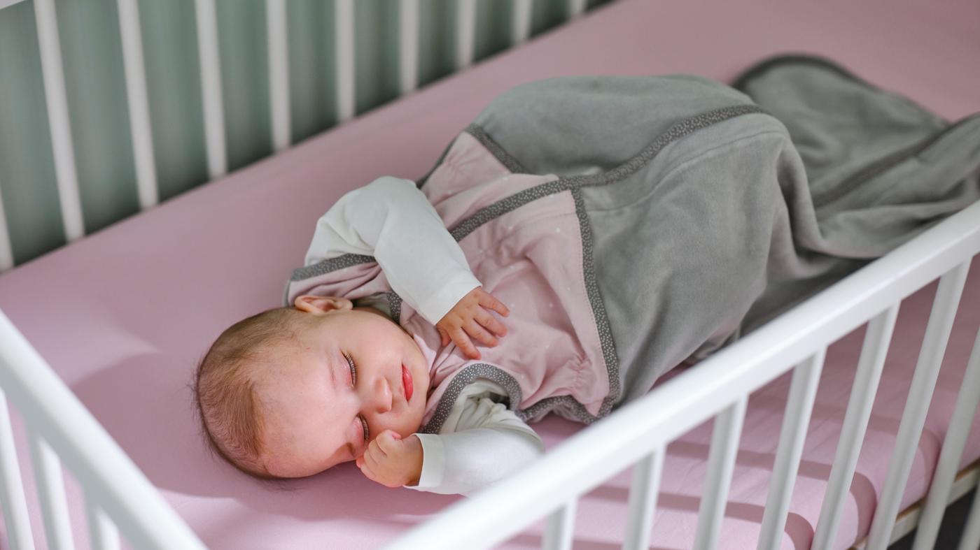 I en vit GULLIVER spjälsäng sover en bebis i en rosa LEN sovpåse på en madrass med ett rosa LEN dra-på-lakan.