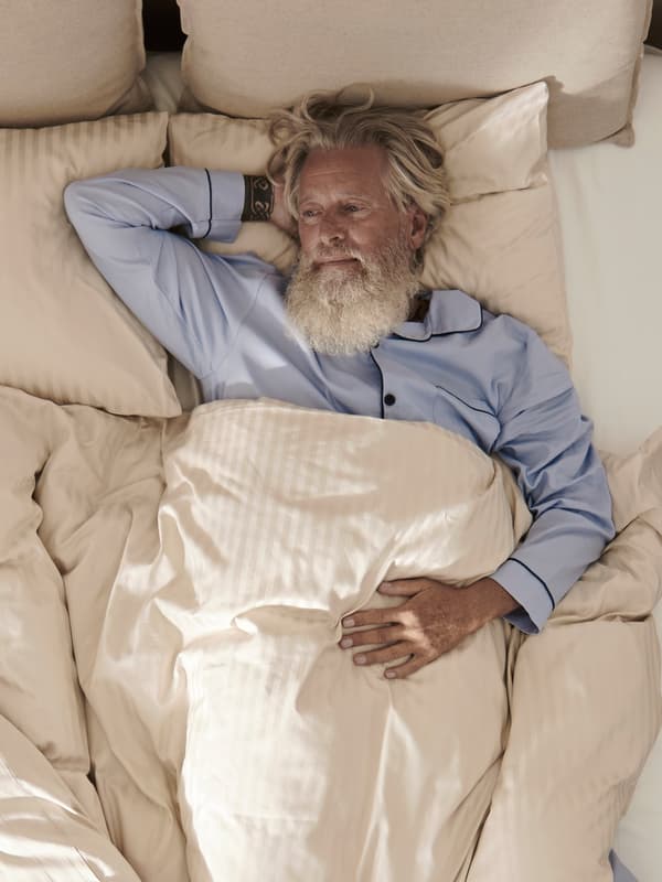 Iemand ligt in een bed met een lichtbeige NATTJASMIN dekbedovertrek en kussenslopen. De persoon staart dromerig in de verte.