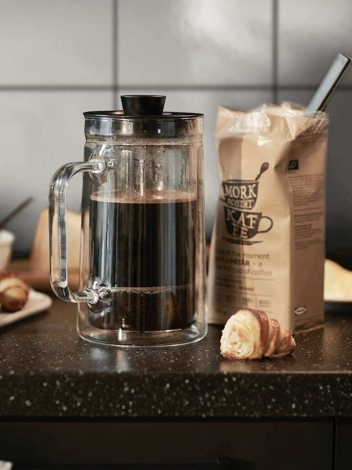 IKEA 365+ konvice na čaj/kávu z čirého skla s nerezovým víkem, naplněná kávou, vedle balení mleté kávy a croissantu na kuchyňské lince.