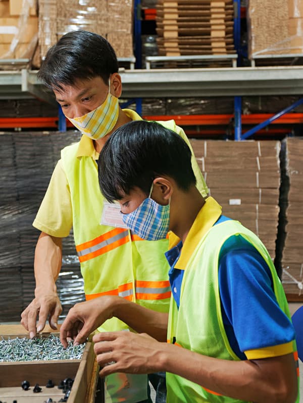 노란색 안전 조끼를 입고 마스크를 쓴 두 명의 IKEA 직원이 공장에서 나사를 검사하고 있습니다.
