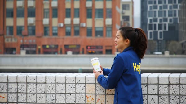 IKEA co‑worker in blue jacket holding a “Hej!” coffee cup, relaxing by a riverside wall with an urban cityscape in the background.