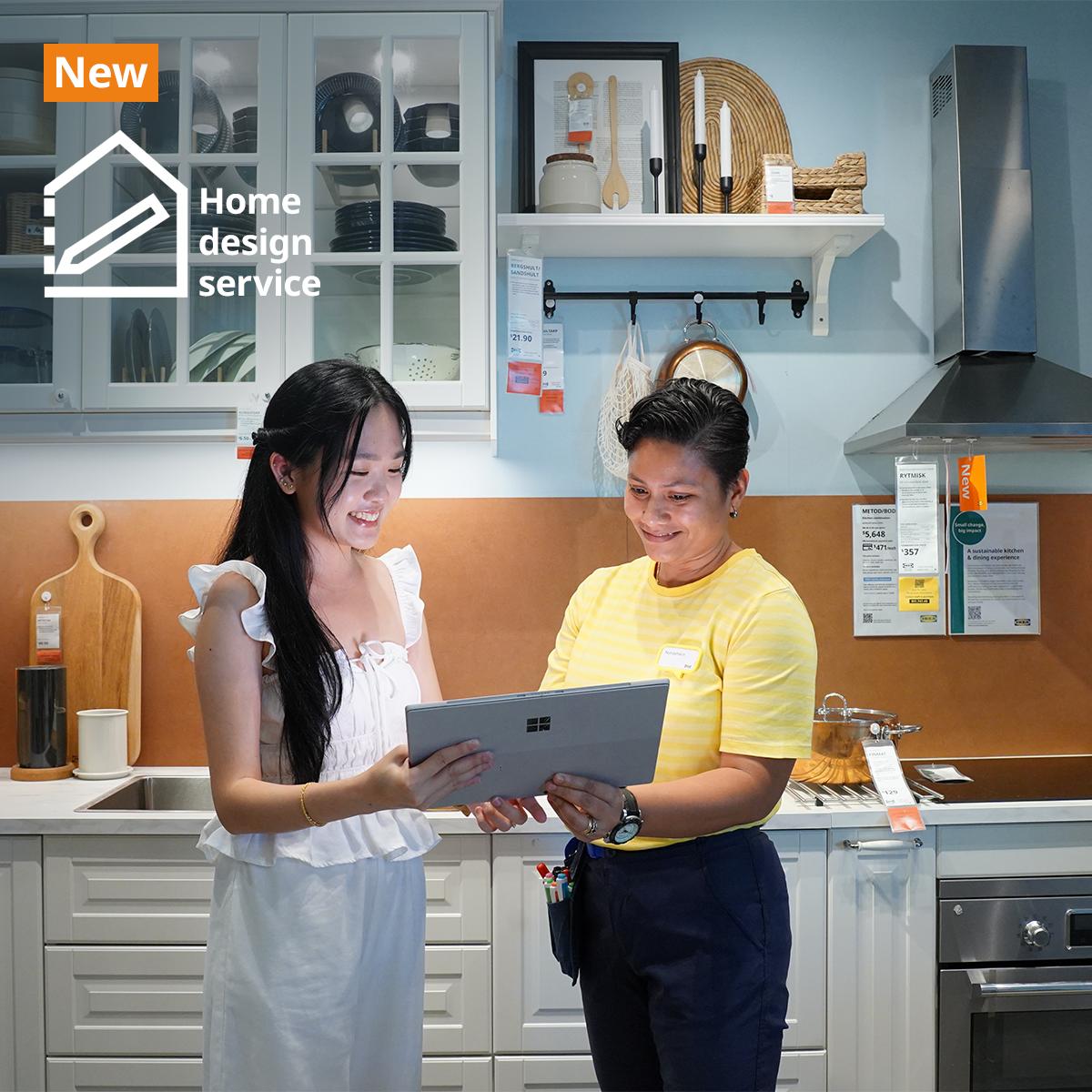 IKEA coworker with a customer in the IKEA kitchen showroom standing together holding a laptop discussing home design. They are standing infront of a METOD kitchen showcase with kitchen cabinets in white fronts and glass doors showing utensils and plates neatly displayed inside. Text on image at the top left hand corner of the image read "New" on an orange background, with a logo and "Home design service" next to it.   