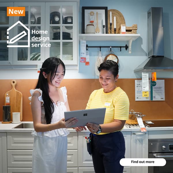 IKEA coworker with a customer in the IKEA kitchen showroom standing together holding a laptop discussing home design. They are standing infront of a METOD kitchen showcase with kitchen cabinets in white fronts and glass doors showing utensils and plates neatly displayed inside. Text on image at the top left hand corner of the image read 