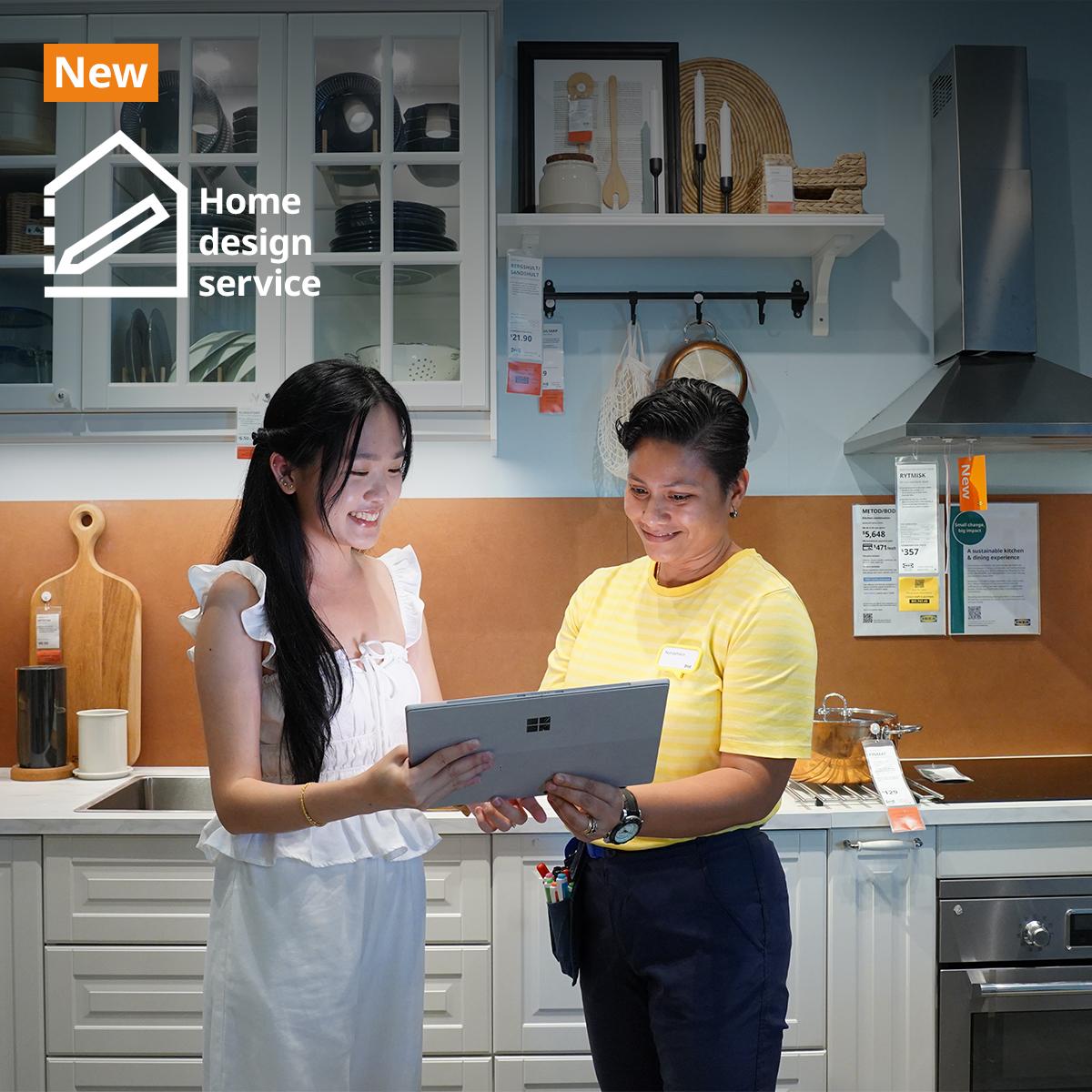 IKEA coworker with a customer in the IKEA kitchen showroom, standing together holding a laptop, discussing home design. They are standing in front of a METOD kitchen showcase with kitchen cabinets in white fronts and glass doors showing utensils and plates neatly displayed inside. Text on image at the top left hand corner of the image read "New" on an orange background, with a logo and "Home design service" next to it.