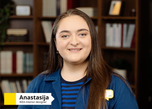 IKEA interior designer wearing a blue uniform, standing in front of a dark wooden bookcase. Text in the bottom-left corner reads: “Anastasija, IKEA interior designer”.						