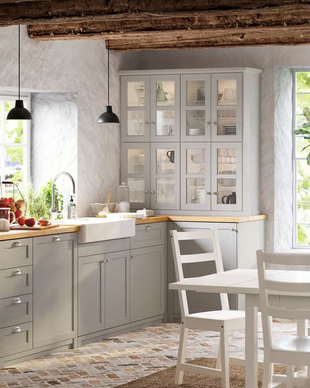 IKEA Küchenlösung mit grauen Fronten, Holzarbeitsplatte, weißem Waschbecken und einer passenden Vitrine die mit Geschirr gefüllt is. Rechts von der Küche ein weißer Esstisch mit passenden weißen Holzstühlen.