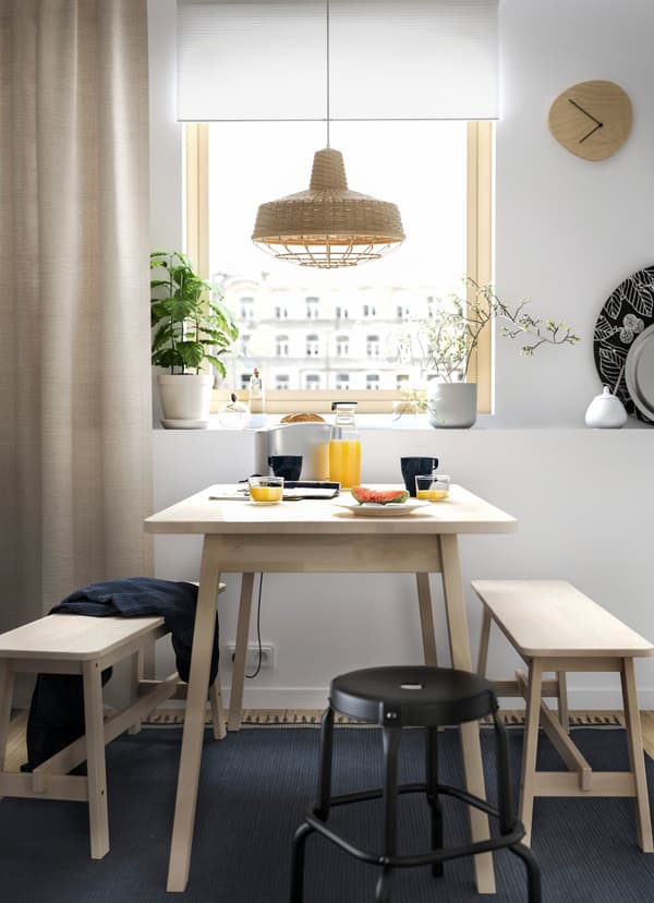 IKEA NORRÅKER light wood bench and one NORRÅKER black stool as dining furniture by a window in the kitchen.