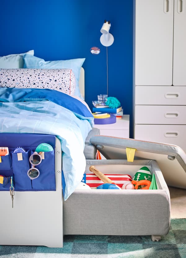 IKEA SLÄKT white bedframe and grey cushioned storage compartment, ajar to reveal sports equipment and toys.