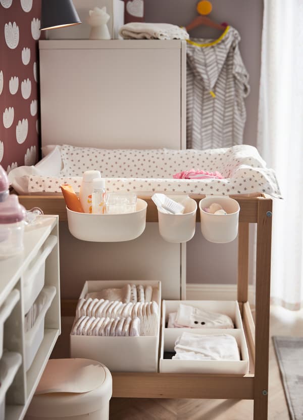 IKEA SNIGLAR baby changing table made of birch with attached ÖNSKLIG white plastic storage baskets holding diapers and baby changing supplies.