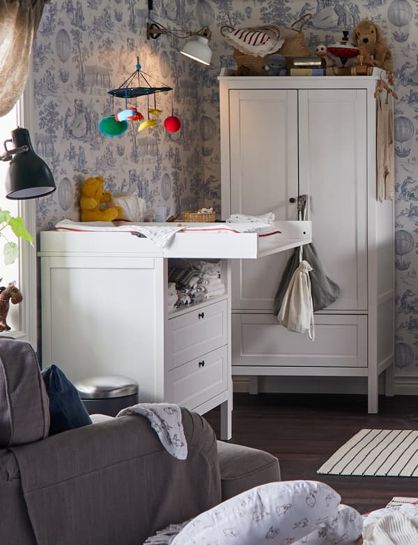 IKEA SUNDVIK changing table in a room with floral wallpaper, wardrobe, armchair, mobile and baby nursing items.