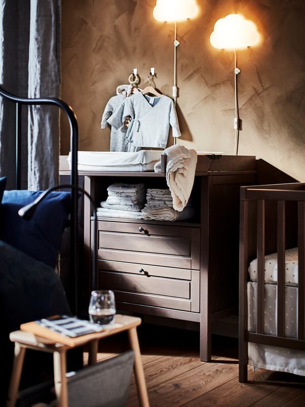 IKEA SUNDVIK changing table in dark wood with baby clothes and baby linens, cot and small side table.