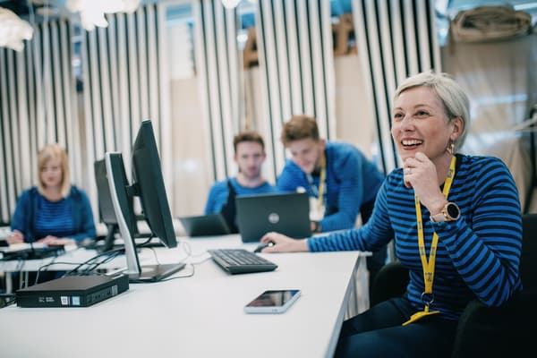 IKEA Team in blauer Arbeitskleidung bei einem produktiven Meeting im Büro, eine Mitarbeiterin lacht freundlich.