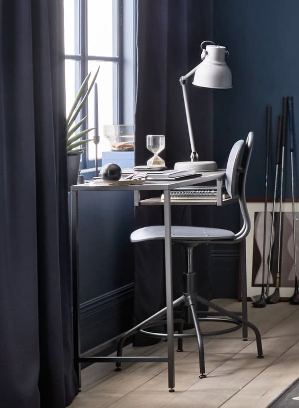 IKEA VITTSJÖ Laptop table in front on a window with a black industrial look chair. On the table there are boxes, some papers and a desk lamp.