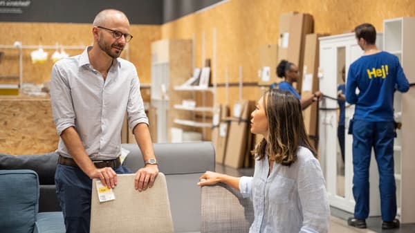 Image of a man and women looking at chairs in the IKEA As-Is department while two IKEA co-workers move a bookcase in the background
