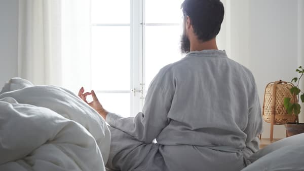 In a calm-looking bedroom, a bearded man sits cross-legged on a bed in a meditation pose, looking out the window.