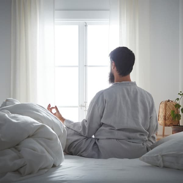 In a calm-looking bedroom, a bearded man sits cross-legged on a bed in a meditation pose, looking out the window.