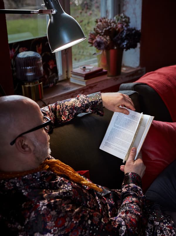 In a cosy living room, a man lounges on a sofa while reading a book. A floor lamp provides lighting as he reads.