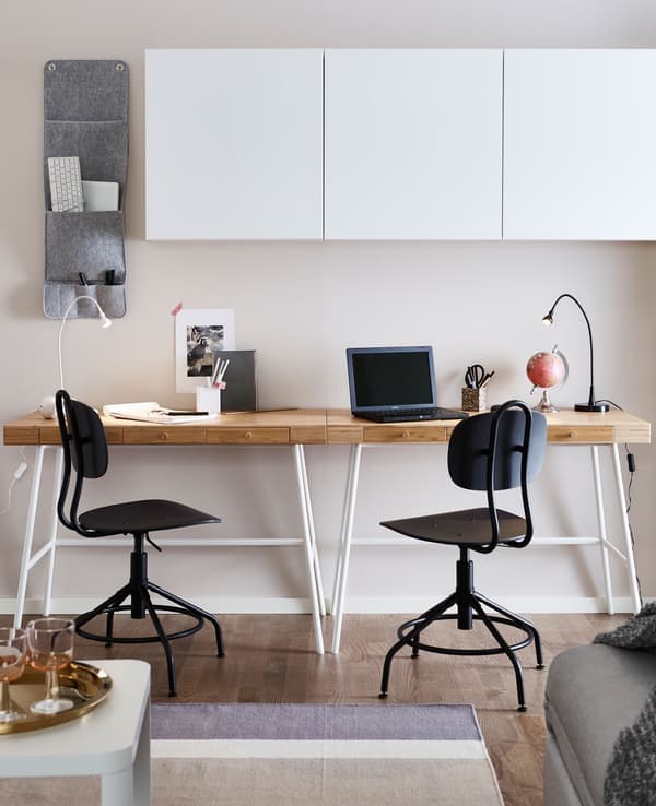 In a living room, two desks sit against a wall, beneath white wall cabinets.
