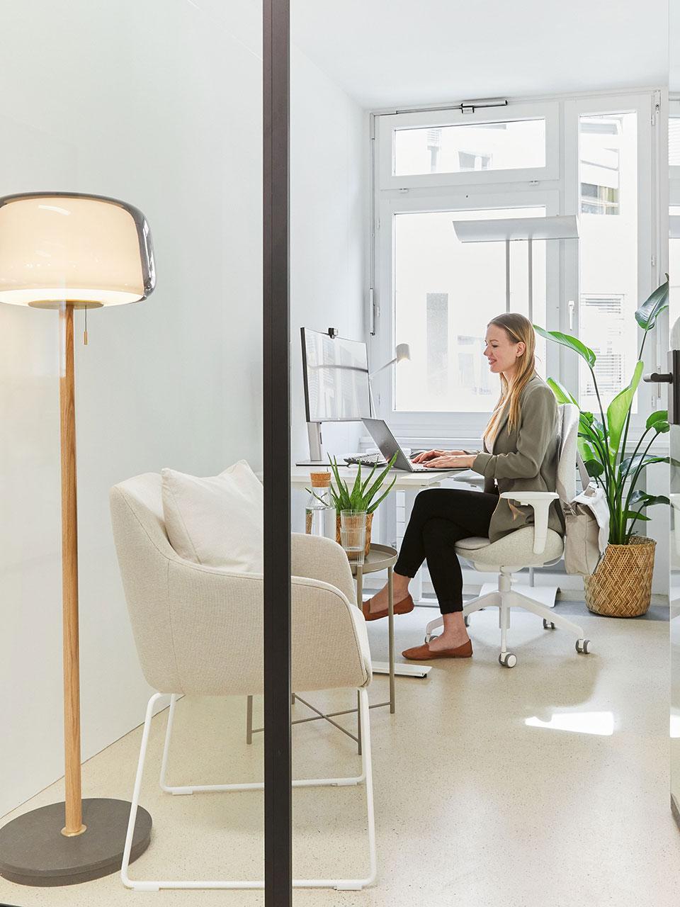 In einem Büro mit Glasfront sitzt eine Frau vor dem Fenster an einem Schreibtisch. Neben ihr steht eine Topfpflanze und davor ein Sessel für Besucher mit Beistelltisch und dahinter eine Stehlampe aus Glas mit einem Fuss aus Holz und Marmor.