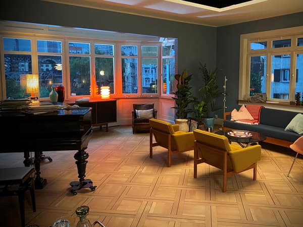 In einem grossen Wohnzimmer mit grosser Fensterfront steht ein Klavier, ein Sideboard, ein Sofa mit Kissen und mehrere Sessel im Retrostil.