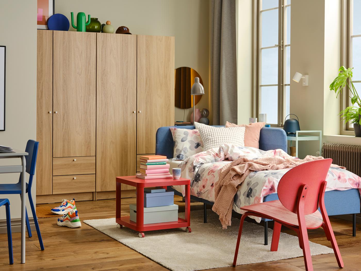 In einem hellen Raum ist ein gepolstertes BLÅKULLEN Bettgestell mit Eckkopfteil in Blau vor einem Kleiderschrank zu sehen. Das Bett ist mit LAPPNYCKLAR Bettwäsche bezogen, davor steht ein FRÖSET Sessel in Rot.