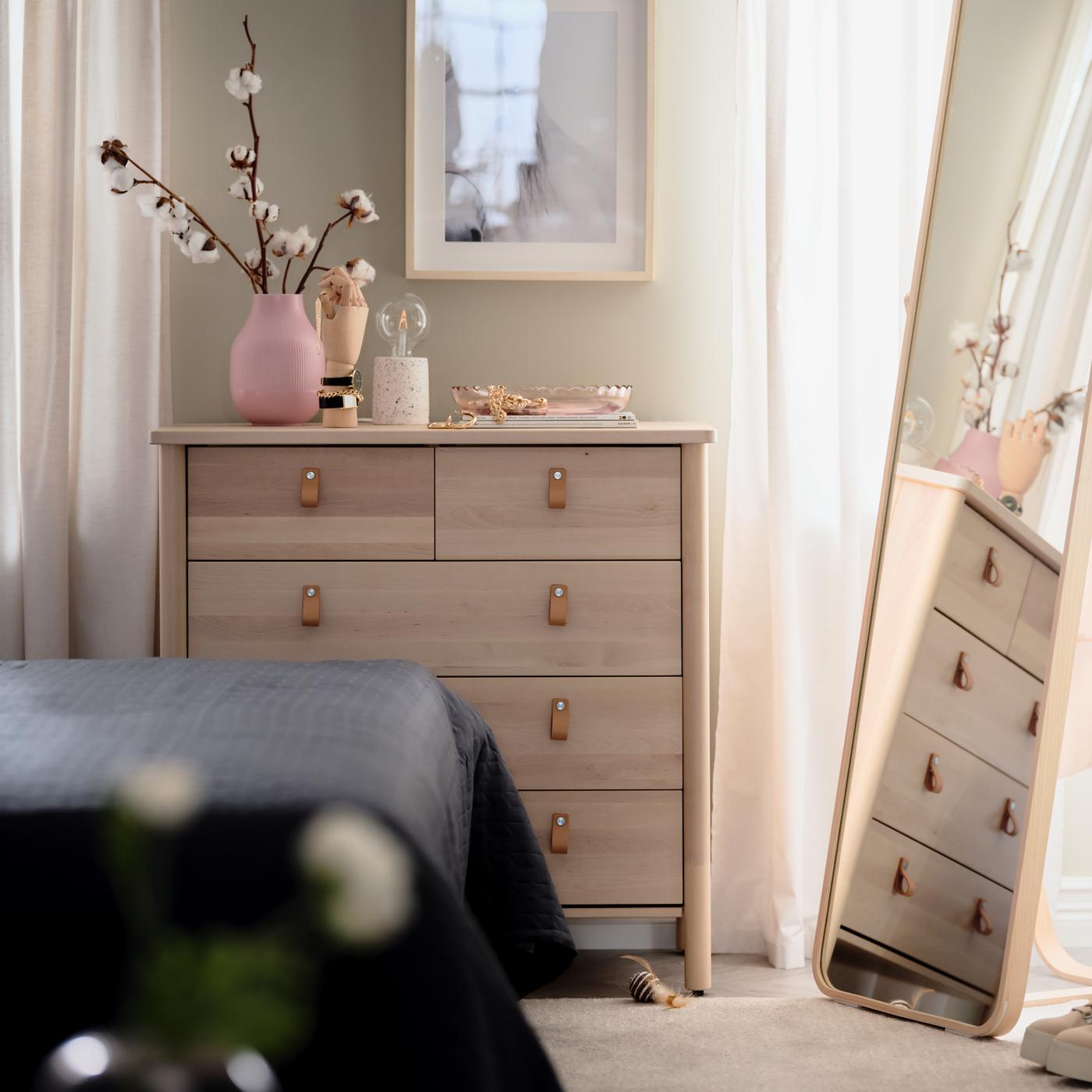 In einem Schlafzimmer ist eine BJÖRKSNÄS Kommode mit 5 Schubladen in Birke zu sehen, auf der sich dekorative Gegenstände befinden. Daneben steht ein IKORNNES Standspiegel aus Esche.