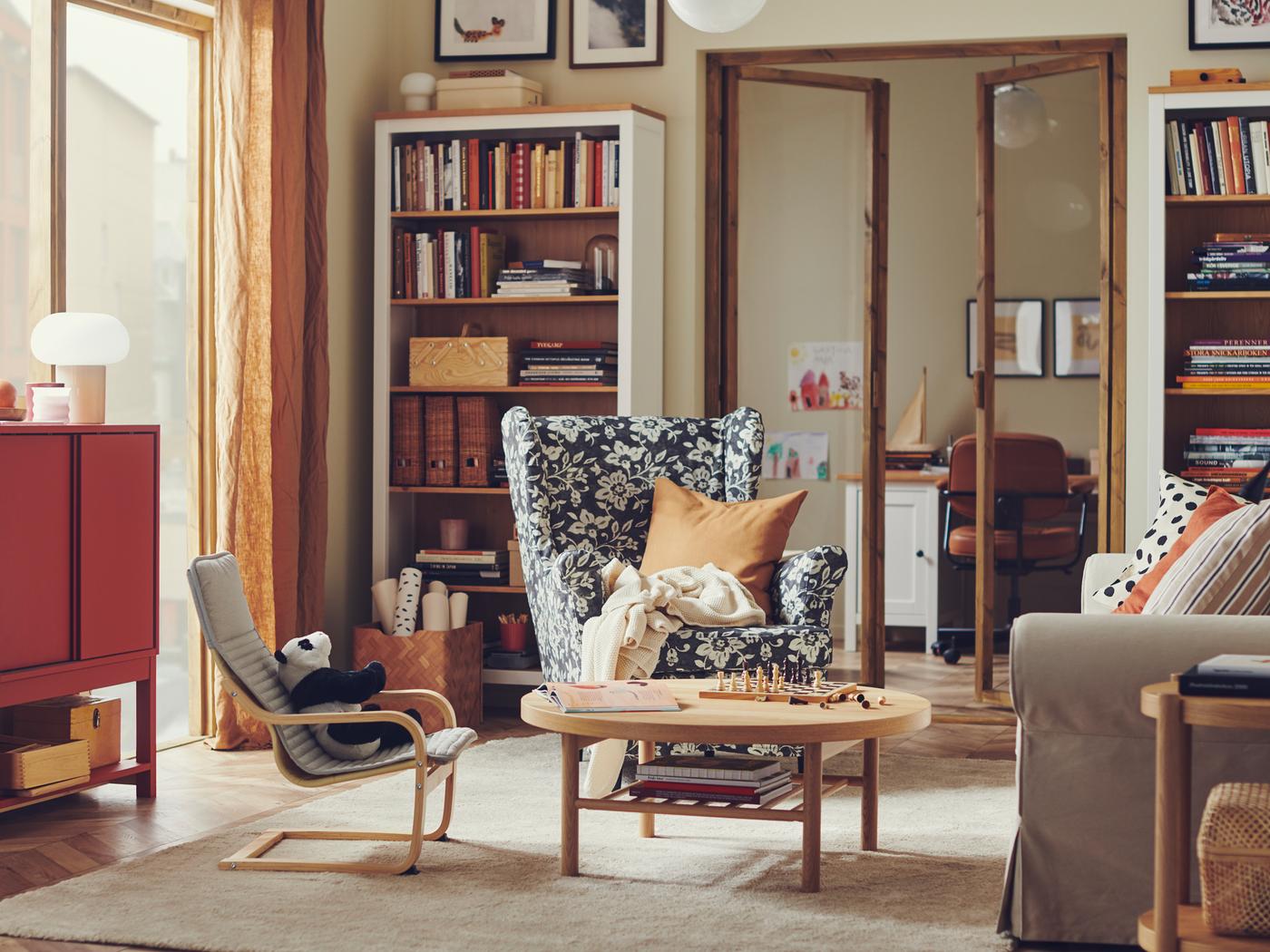In einem Wohnzimmer in einem warmen traditionellen Stil steht ein STRANDMON Ohrensessel vor einem HEMNES Bücherregal voller Bücher und Gegenstände.