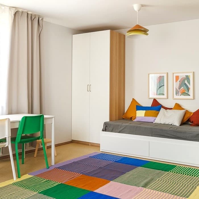 In the family room, there is a bed with multicoloured cushions, a two-door wardrobe, a desk/dining table and a colourful rug on the floor.