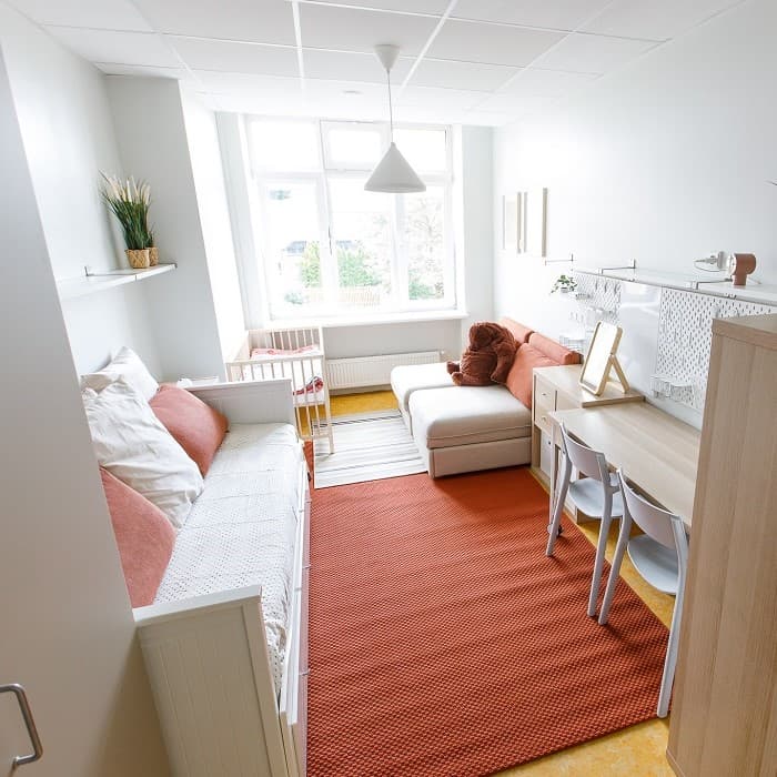 In the family room, there is a desk, a daybed, a baby cot and two pull-out armchairs, with an orange rug laid on the floor.