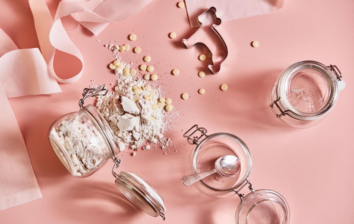 Ingrédients pour réaliser des biscuits, bocaux et emporte-pièces, sur un fond rose, vus d’en haut.