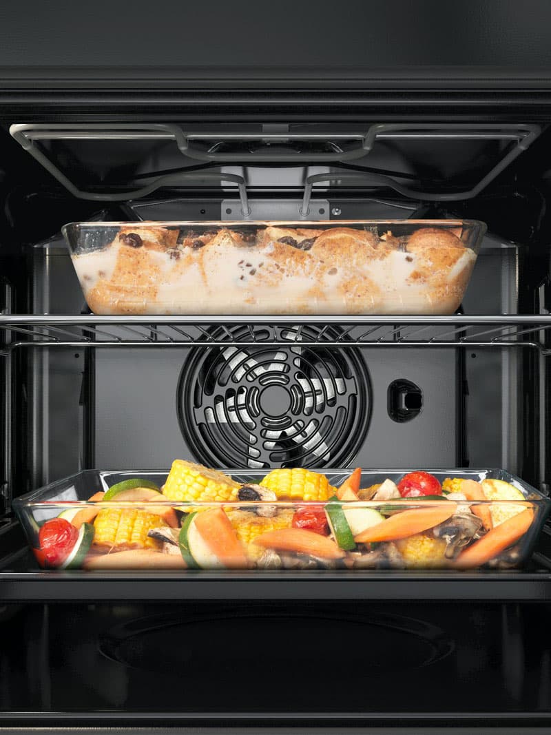 Innenansicht eines Backofens mit zwei Blechen: Oben ein gebackenes Gericht, unten verschiedenes Gemüse wie Paprika und Zucchini, zum Rösten angerichtet.