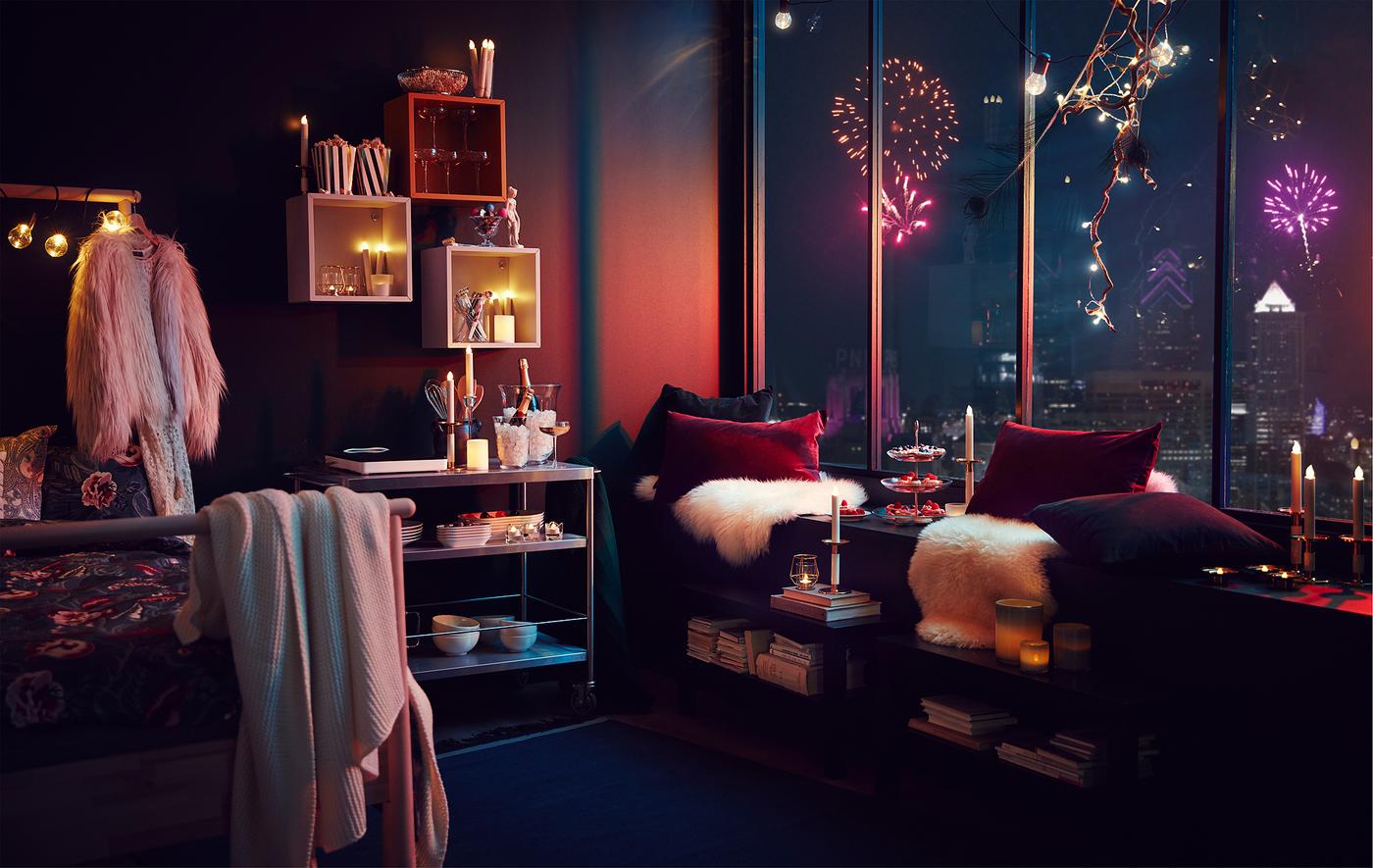 Intérieur d’appartement avec décorations et éclairage à la fête ; feux d’artifice à l’horizon d’une ville visibles à travers de hautes fenêtres.