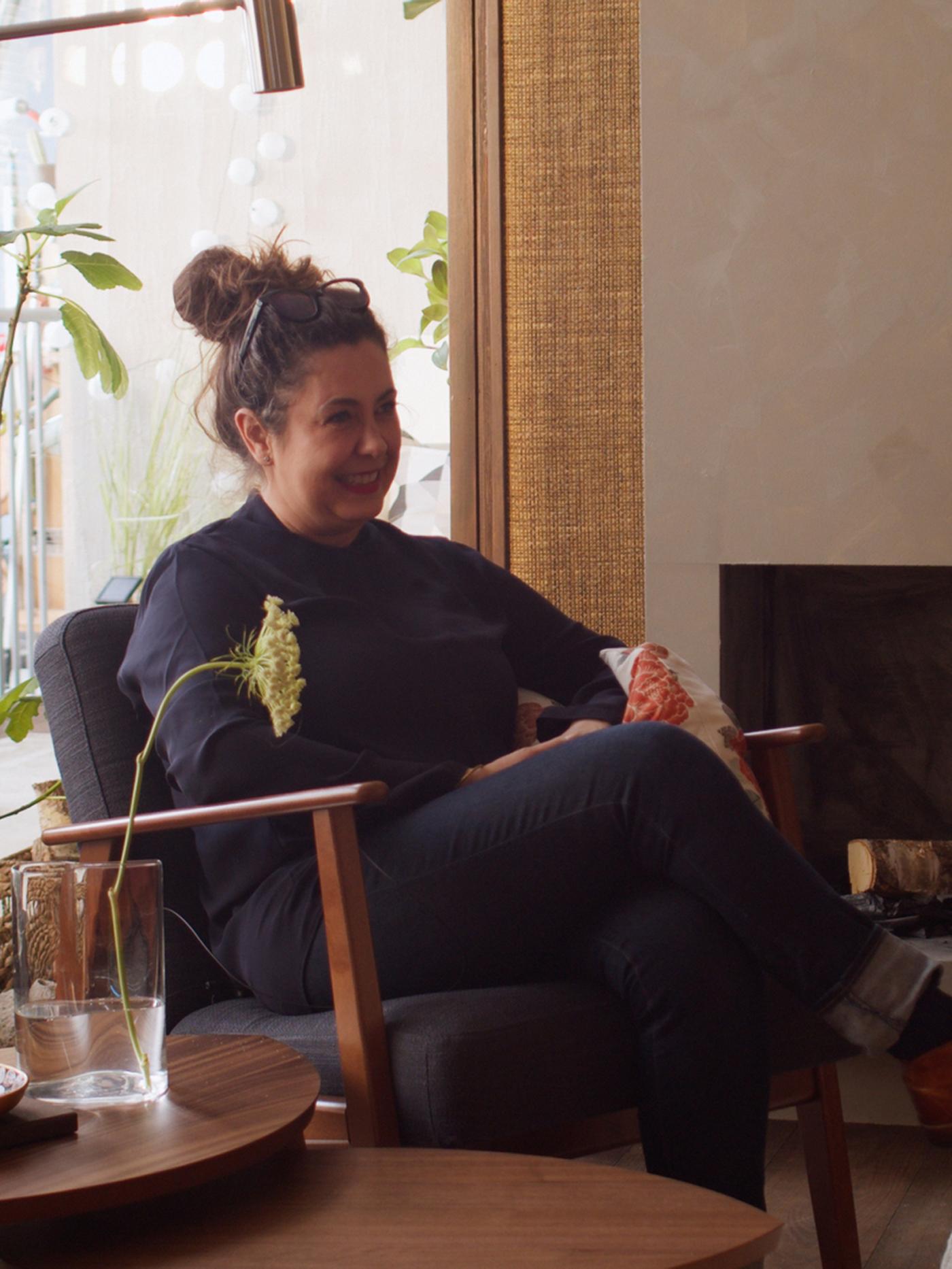 Interior designer Chiara Effroi Lutteri, sitting in an armchair in front of a window with tables with a flower in a vase.