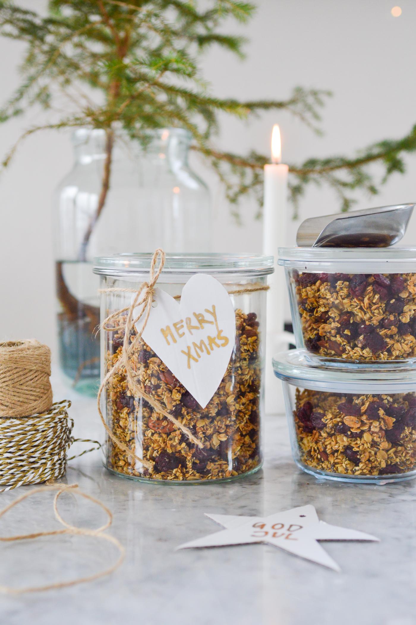 Jars filled with muesli and labelled 'Merry Xmas' stand on a marble slab, surrounded by string, a white candle and a small tree, creating a festive, homemade atmosphere.